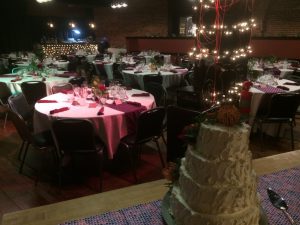 Tables are set up with tablecloths and silverware behind a tall, white wedding cake in SPARK Museum's Powerhouse event center.